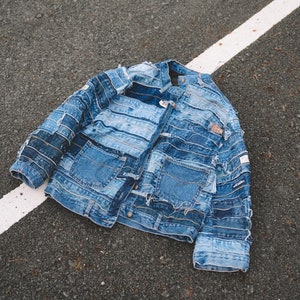 Puede incluir: Una chaqueta vaquera azul hecha de múltiples capas de denim reciclado. La chaqueta tiene un diseño clásico con cierre de botones y dos bolsillos.