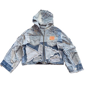 Op de afbeelding: Een blauwe denimjas met een capuchon, gemaakt van patchwork stukken denim. De jas heeft een ritssluiting en rafelige randen.