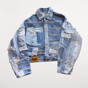 Peut inclure: Une veste en jean bleue composée de multiples panneaux en patchwork de différentes nuances de bleu. La veste présente un col, une fermeture boutonnée sur le devant, deux poches poitrine et des bords effilochés. Un écusson en cuir marron avec le texte NIAT. est visible.