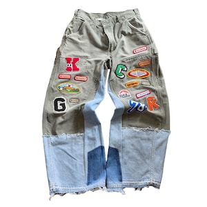 May include: Olive green and light blue denim pants featuring a patchwork design with frayed hems. The pants are decorated with various colorful embroidered patches, including letters, shapes, and text such as Las Vegas and Recruiter.