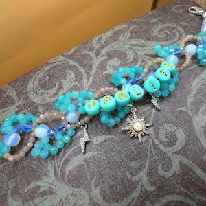 Puede incluir: Una pulsera de cuentas con la palabra "OPAL" escrita con cuentas de letras turquesas. La pulsera presenta varias cuentas azules y transparentes, dijes de rayo plateado y un dije de sol. También hay un pequeño dije de corona rosa.