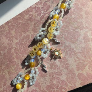 May include: A handmade beaded bracelet with the word "OPALITE" spelled out in yellow letter beads. The bracelet features white and yellow beads, a sun charm, and a lightning bolt charm. The bracelet is on a pink floral background.