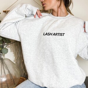 May include: A gray sweatshirt with the text "LASH ARTIST" printed in black on the front.