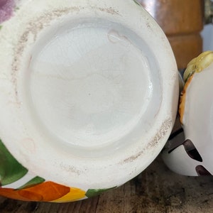 Puede incluir: La parte inferior de una jarra de cer&aacute;mica blanca con un dise&ntilde;o floral colorido. La jarra tiene una base redonda y una abertura ancha. El dise&ntilde;o floral incluye flores naranjas, amarillas, verdes y moradas.