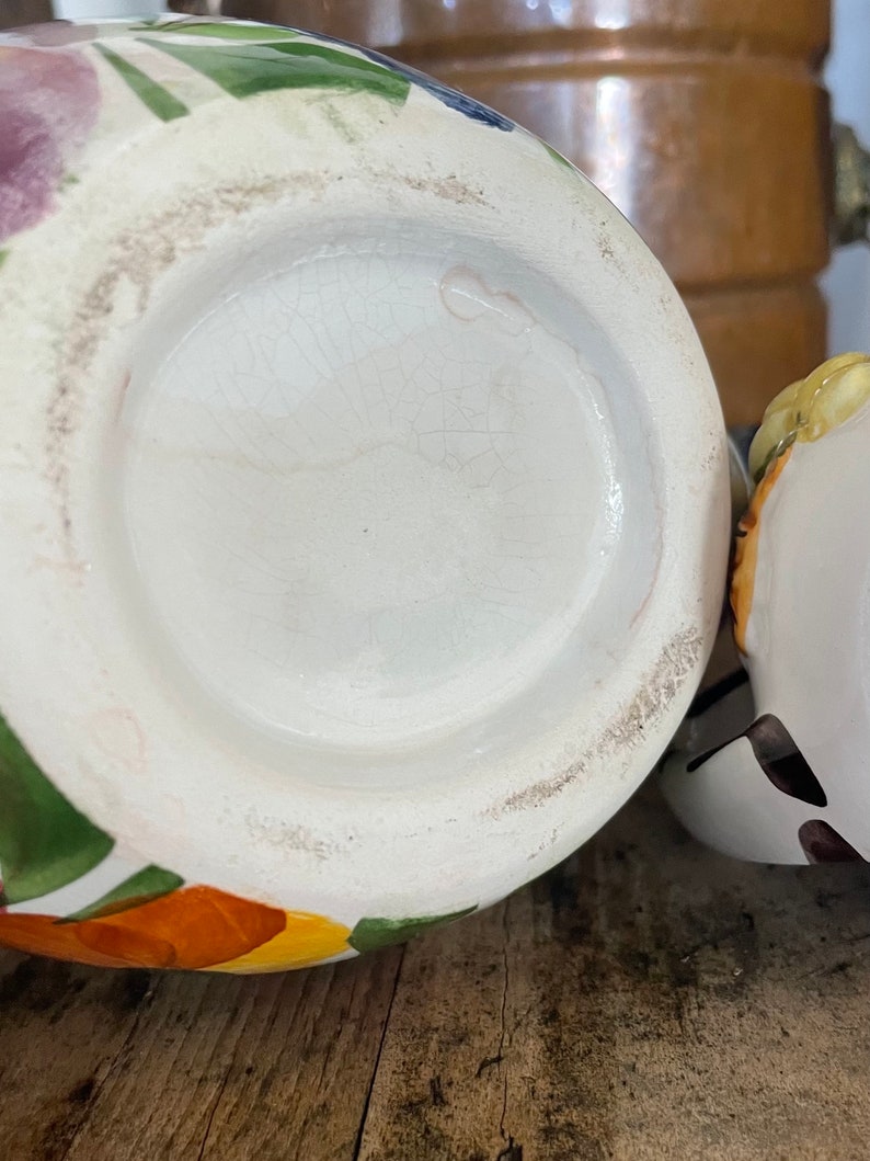 Puede incluir: La parte inferior de una jarra de cer&aacute;mica blanca con un dise&ntilde;o floral colorido. La jarra tiene una base redonda y una abertura ancha. El dise&ntilde;o floral incluye flores naranjas, amarillas, verdes y moradas.