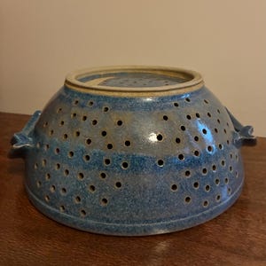 K&ouml;nnte beinhalten: Handgefertigte blaue Keramik-Abtropfschale mit zahlreichen kleinen L&ouml;chern zum Ablaufen. Die Abtropfschale hat eine breite, abgerundete Form mit zwei kleinen Griffen auf beiden Seiten. Der Rand ist in einem helleren Blauton gehalten.