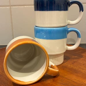 May include: Three ceramic mugs stacked on a wooden surface. The mugs have a two-tone design with bands of blue, white, and orange. One mug is on its side, showing the inside. The mugs have curved handles.