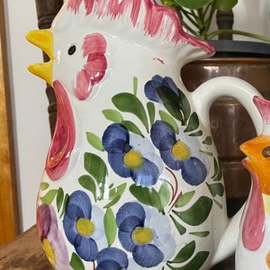 Puede incluir: Jarra de cer&aacute;mica blanca con forma de gallo con cresta y barbilla rojas. La jarra est&aacute; decorada con flores azules y amarillas y hojas verdes.