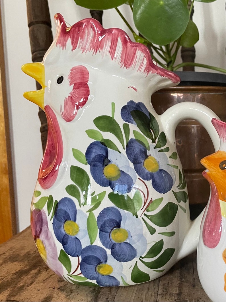 Puede incluir: Jarra de cer&aacute;mica blanca con forma de gallo con cresta y barbilla rojas. La jarra est&aacute; decorada con flores azules y amarillas y hojas verdes.