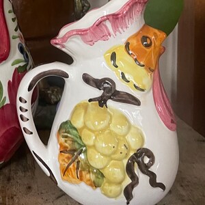 Puede incluir: Jarra de cer&aacute;mica blanca con un dise&ntilde;o de gallo. El gallo tiene una cresta y barbilla rosadas, plumas amarillas y naranjas y un pico marr&oacute;n. La jarra tambi&eacute;n presenta un dise&ntilde;o pintado de un racimo de uvas con hojas verdes y una vid marr&oacute;n.