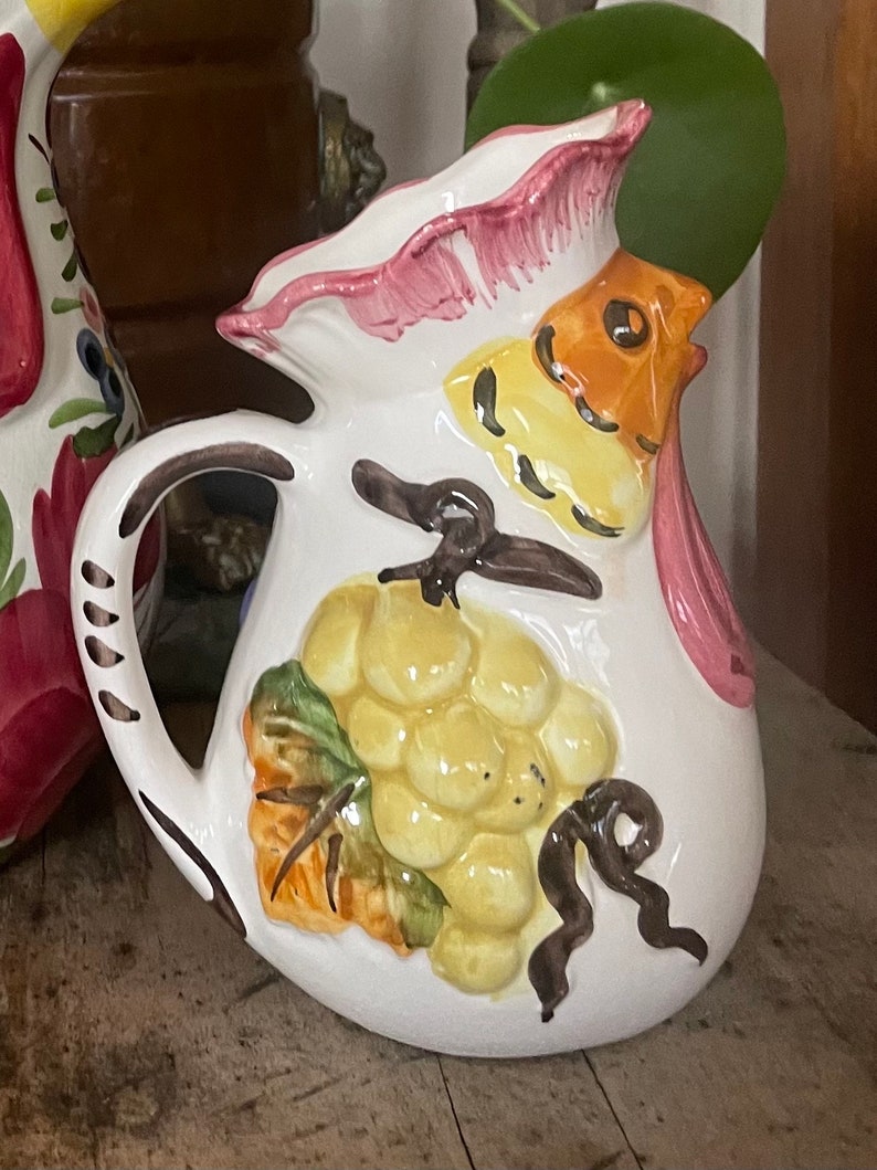 Puede incluir: Jarra de cer&aacute;mica blanca con un dise&ntilde;o de gallo. El gallo tiene una cresta y barbilla rosadas, plumas amarillas y naranjas y un pico marr&oacute;n. La jarra tambi&eacute;n presenta un dise&ntilde;o pintado de un racimo de uvas con hojas verdes y una vid marr&oacute;n.
