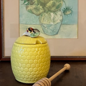May include: A yellow ceramic honey pot with a honeycomb design, adorned with a bird figurine. A wooden honey dipper is placed next to the pot. The background shows a framed painting of sunflowers in a vase.