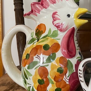 Puede incluir: Una jarra de cer&aacute;mica blanca con forma de gallo con cresta rosa y pico amarillo. La jarra est&aacute; decorada con flores naranjas, amarillas y verdes.