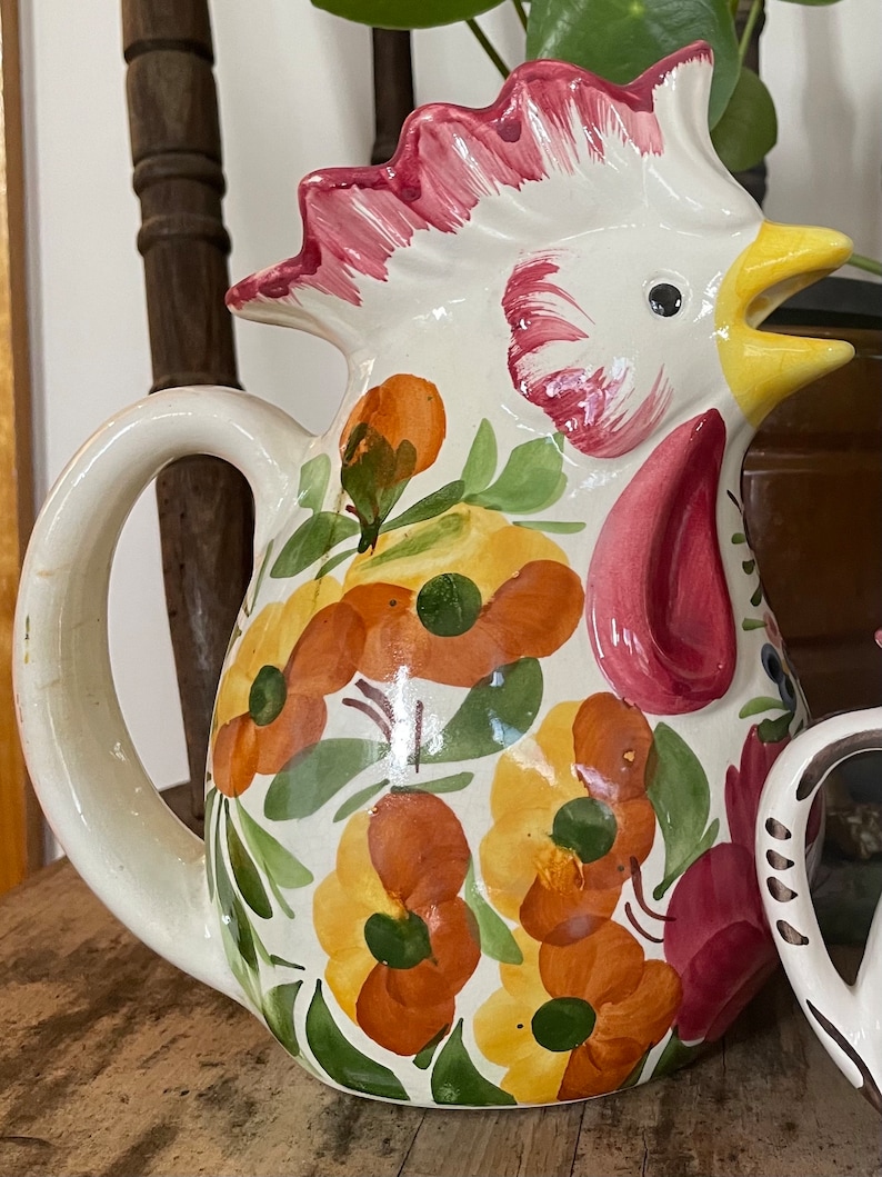 Puede incluir: Una jarra de cer&aacute;mica blanca con forma de gallo con cresta rosa y pico amarillo. La jarra est&aacute; decorada con flores naranjas, amarillas y verdes.