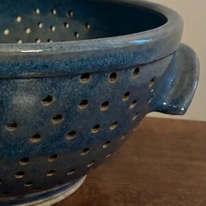 K&ouml;nnte beinhalten: Nahaufnahme einer blauen Keramik-Gem&uuml;sesp&uuml;le mit zahlreichen kleinen L&ouml;chern. Die Gem&uuml;sesp&uuml;le hat eine runde Form mit zwei Griffen und einem leicht ausgestellten Rand. Die Keramik hat eine gesprenkelte Textur.