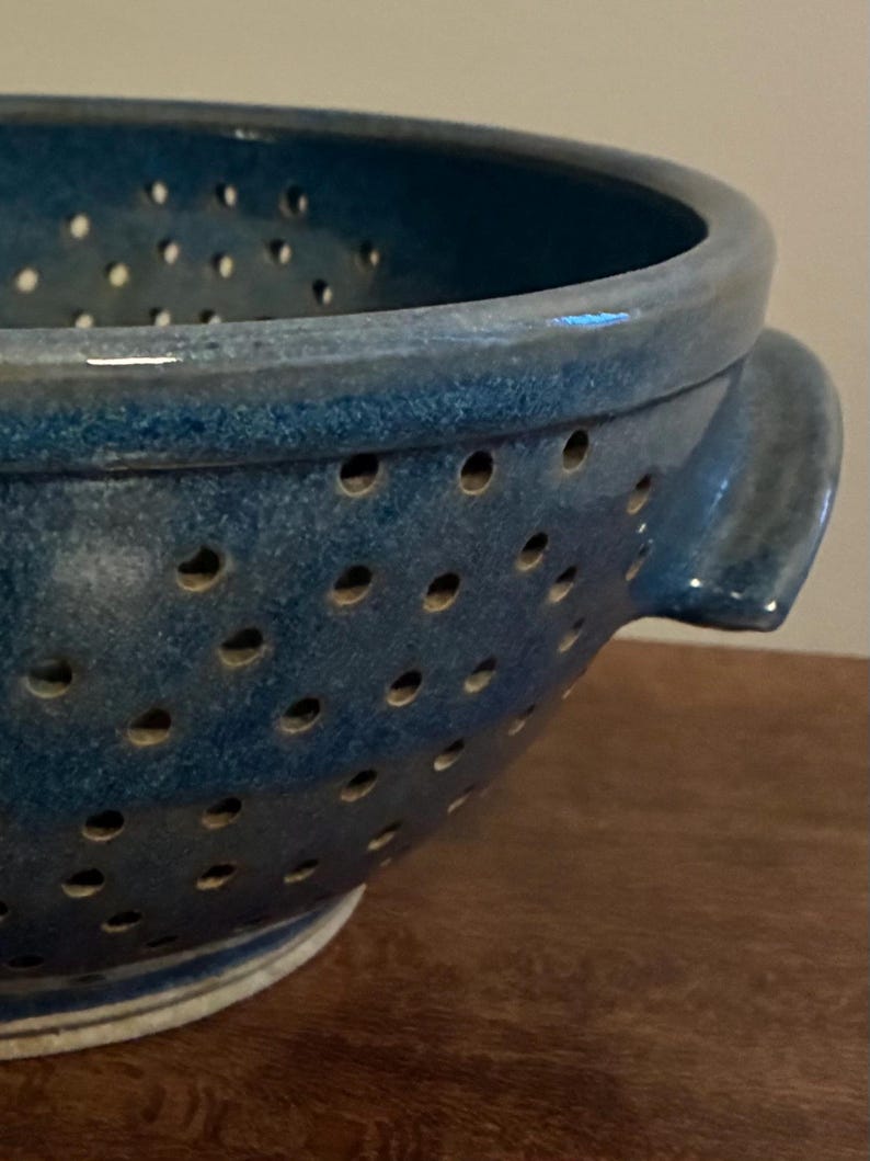 K&ouml;nnte beinhalten: Nahaufnahme einer blauen Keramik-Gem&uuml;sesp&uuml;le mit zahlreichen kleinen L&ouml;chern. Die Gem&uuml;sesp&uuml;le hat eine runde Form mit zwei Griffen und einem leicht ausgestellten Rand. Die Keramik hat eine gesprenkelte Textur.
