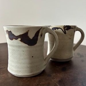 May include: Two ceramic mugs with brown glaze designs on a light beige background. The mugs have a rounded shape and a handle on the side.