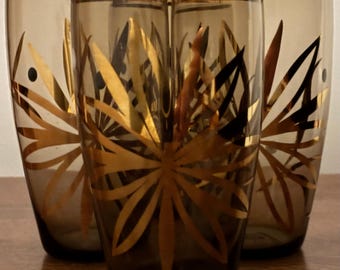 Puede incluir: Un juego de tres vasos de cristal ahumado con un patrón de estrella o floral en tono dorado en el exterior. Cada vaso tiene una forma cilíndrica cónica y lisa, con un borde dorado metálico en la parte superior, creando un conjunto uniforme.