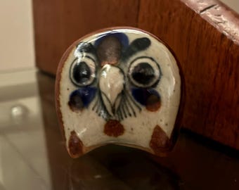 Puede incluir: Figura de búho de cerámica con base blanca, detalles azules, marrones y negros. El búho tiene ojos grandes y redondos y un pico pequeño. La figura es un objeto decorativo.