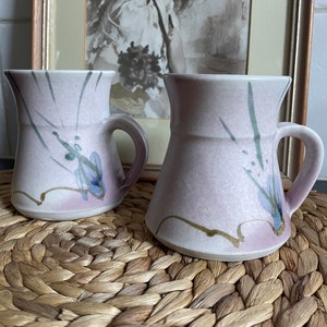 May include: Two ceramic mugs with a light pink glaze and abstract blue and green brushstrokes. The mugs have a rounded body and a handle on the side.