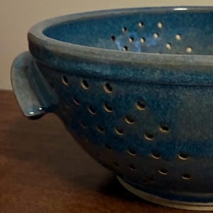 K&ouml;nnte beinhalten: Nahaufnahme einer handgefertigten, blauen Keramik-Gem&uuml;sesp&uuml;le. Die Gem&uuml;sesp&uuml;le hat eine runde Form mit zahlreichen kleinen L&ouml;chern und zwei Griffen. Die Glasur hat ein strukturiertes Aussehen.