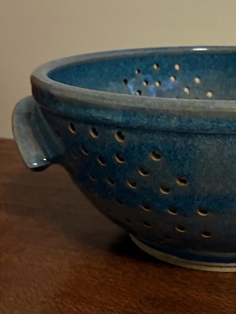 K&ouml;nnte beinhalten: Nahaufnahme einer handgefertigten, blauen Keramik-Gem&uuml;sesp&uuml;le. Die Gem&uuml;sesp&uuml;le hat eine runde Form mit zahlreichen kleinen L&ouml;chern und zwei Griffen. Die Glasur hat ein strukturiertes Aussehen.