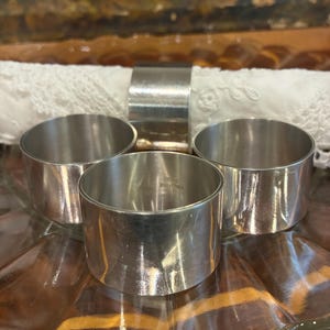May include: A collection of four silver napkin rings. Three rings are arranged in a semi-circle, with a fourth ring behind them. The rings are on a clear, scalloped glass plate. The background includes a white lace napkin and a painting.