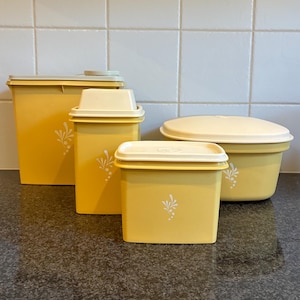 May include: A collection of four vintage yellow food storage containers with cream-coloured lids. Each container features a white floral design. The containers vary in size and shape, including a tall rectangular container, a square container, and a round container.