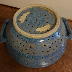 K&ouml;nnte beinhalten: Ein blauer Keramik-Gem&uuml;sesieb mit zahlreichen kleinen L&ouml;chern zum Ablaufen. Das Sieb hat einen breiten Rand und zwei Griffe. Die Farbe ist ein gesprenkeltes Blau mit einigen helleren Streifen. Das Sieb steht auf einer Holzoberfl&auml;che.