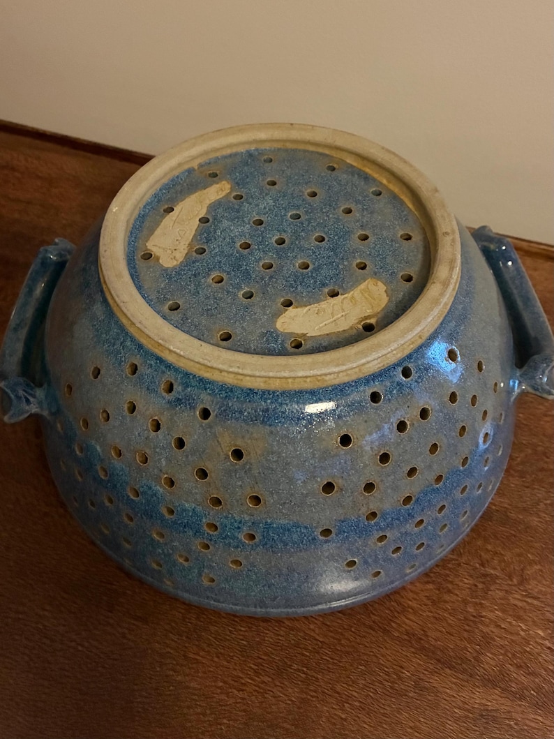 K&ouml;nnte beinhalten: Ein blauer Keramik-Gem&uuml;sesieb mit zahlreichen kleinen L&ouml;chern zum Ablaufen. Das Sieb hat einen breiten Rand und zwei Griffe. Die Farbe ist ein gesprenkeltes Blau mit einigen helleren Streifen. Das Sieb steht auf einer Holzoberfl&auml;che.