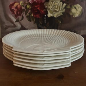 May include: A stack of six white ceramic plates with a fan-like design radiating from the centre. The plates have a unique, curved handle on one side. The plates are stacked on a wooden surface.