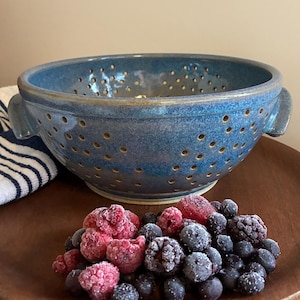 May include: A blue ceramic colander with many small holes and two handles. A wooden plate holds a mix of frozen berries, including raspberries, blueberries, and blackberries. A striped towel is partially visible.
