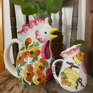 Puede incluir: Dos jarras de cer&aacute;mica, una con forma de gallo con dise&ntilde;os florales en tonos naranja, amarillo, verde y rosa, y la otra con forma de p&aacute;jaro con un racimo de uvas pintado.