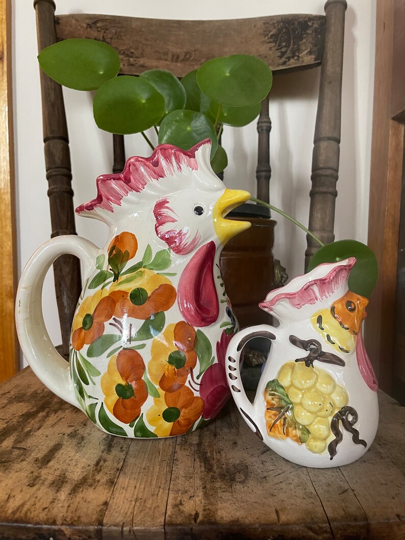 Puede incluir: Dos jarras de cer&aacute;mica, una con forma de gallo con dise&ntilde;os florales en tonos naranja, amarillo, verde y rosa, y la otra con forma de p&aacute;jaro con un racimo de uvas pintado.