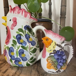 Puede incluir: Dos jarras de cer&aacute;mica con forma de gallo con dise&ntilde;os florales y de uvas. La jarra m&aacute;s grande es blanca con flores azules y hojas verdes. La jarra m&aacute;s peque&ntilde;a es blanca con un gallo amarillo, hojas verdes y uvas moradas.