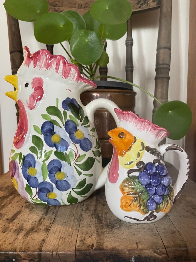 Puede incluir: Dos jarras de cer&aacute;mica con forma de gallo con dise&ntilde;os florales y de uvas. La jarra m&aacute;s grande es blanca con flores azules y hojas verdes. La jarra m&aacute;s peque&ntilde;a es blanca con un gallo amarillo, hojas verdes y uvas moradas.