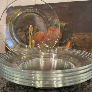May include: A stack of clear glass dinner plates with a slightly rippled edge. The plates are stacked on a dark gray surface.