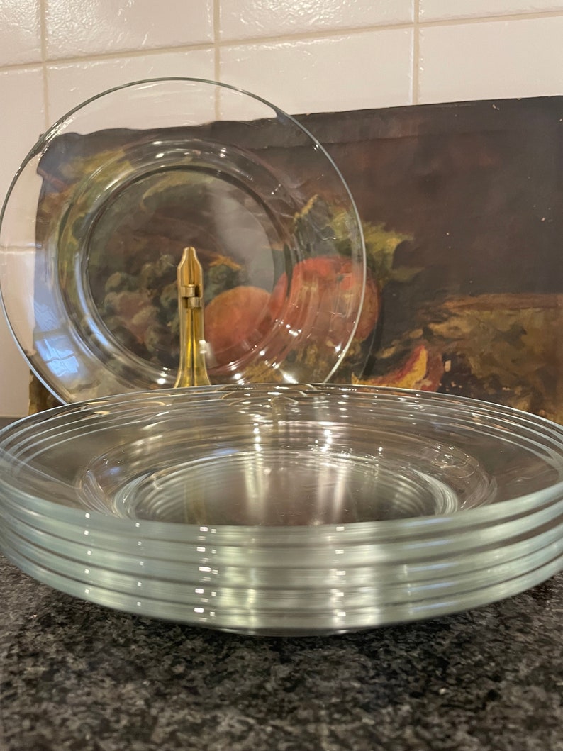 May include: A stack of clear glass dinner plates with a slightly rippled edge. The plates are stacked on a dark gray surface.