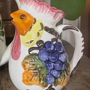 Puede incluir: Una jarra de cer&aacute;mica blanca con un dise&ntilde;o de gallo. El gallo est&aacute; pintado en tonos de naranja, amarillo y rosa. La jarra tambi&eacute;n presenta un racimo de uvas moradas y hojas verdes.
