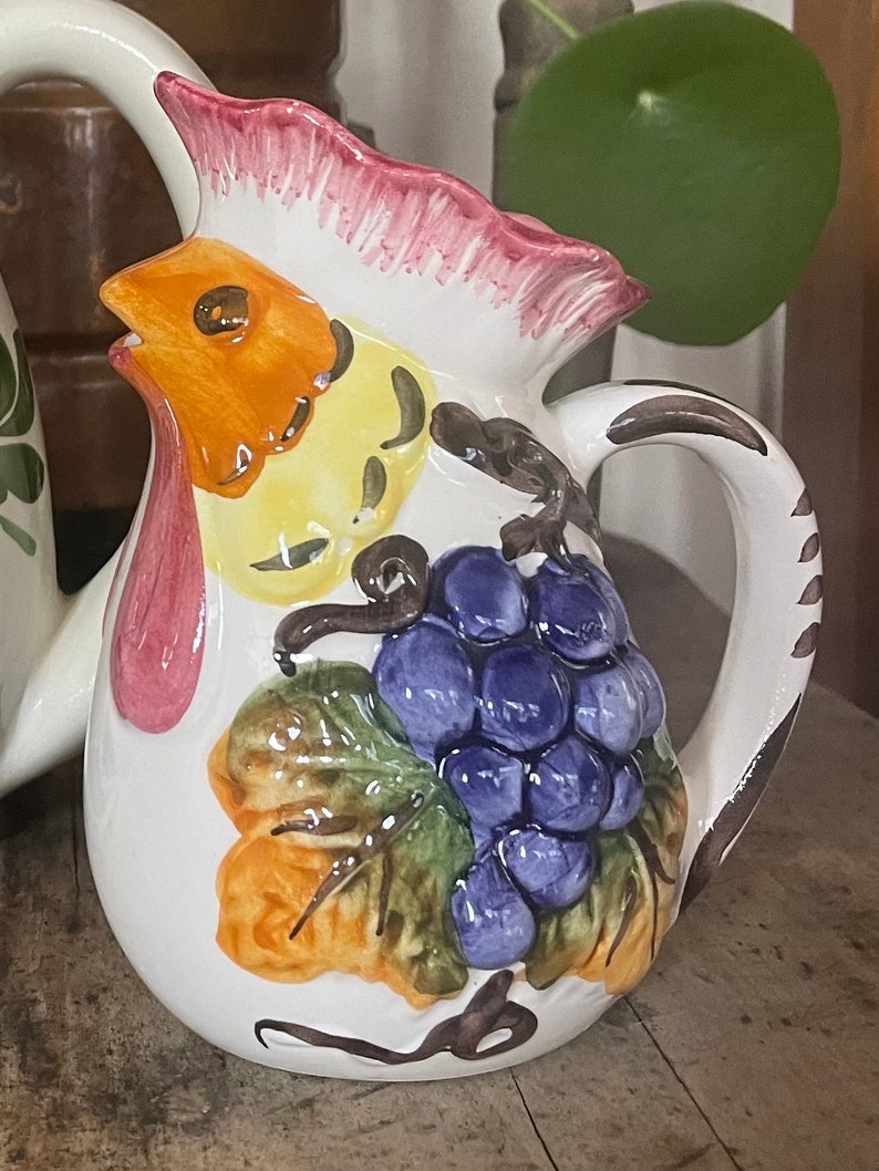 Puede incluir: Una jarra de cer&aacute;mica blanca con un dise&ntilde;o de gallo. El gallo est&aacute; pintado en tonos de naranja, amarillo y rosa. La jarra tambi&eacute;n presenta un racimo de uvas moradas y hojas verdes.