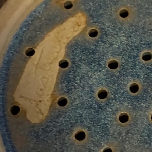 K&ouml;nnte beinhalten: Nahaufnahme einer Keramik-Gem&uuml;sesch&uuml;ssel mit blau gesprenkeltem Innenraum und zahlreichen kleinen L&ouml;chern. Ein Teil des Randes der Gem&uuml;sesch&uuml;ssel ist sichtbar, zusammen mit einem hellfarbigen, unregelm&auml;&szlig;igen Fleck mit verblasstem Text.