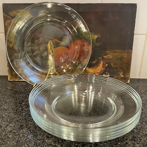 May include: A stack of clear glass dinner plates. The plates are round and have a slightly raised rim. The plates are stacked on a black countertop.