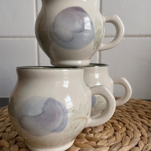May include: Three white ceramic mugs with a light blue and purple floral design. The mugs have a rounded body and a handle.