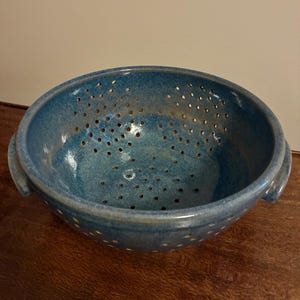 K&ouml;nnte beinhalten: Eine handgefertigte, blaue Keramik-Salatsch&uuml;ssel mit gesprenkelter Glasur. Die Sch&uuml;ssel hat zahlreiche kleine L&ouml;cher zum Ablaufen und zwei kleine Griffe. Die Salatsch&uuml;ssel steht auf einer Holzoberfl&auml;che.