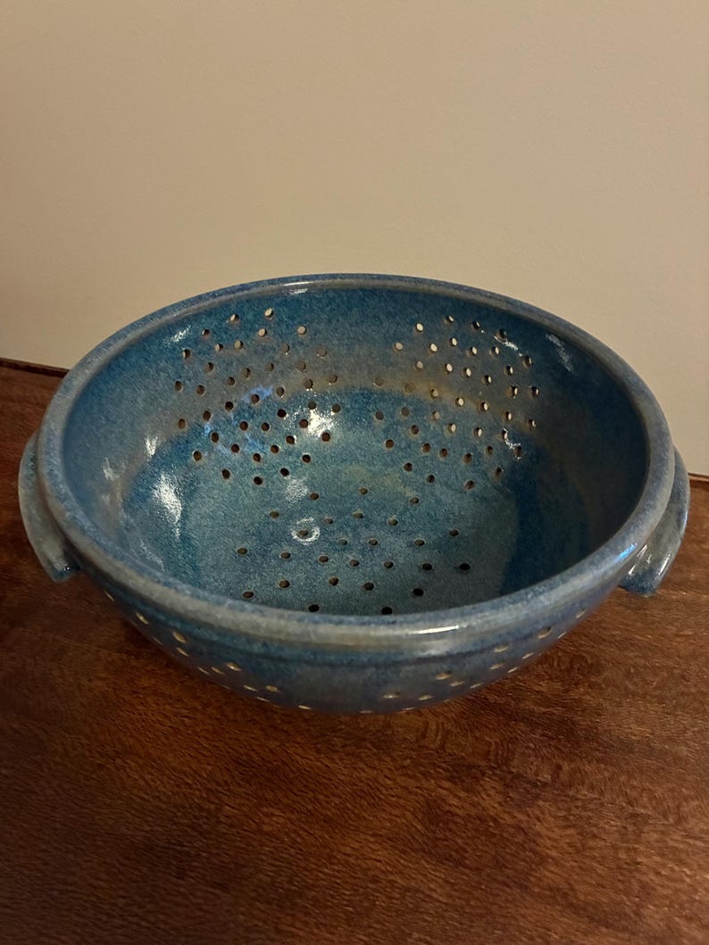 K&ouml;nnte beinhalten: Eine handgefertigte, blaue Keramik-Salatsch&uuml;ssel mit gesprenkelter Glasur. Die Sch&uuml;ssel hat zahlreiche kleine L&ouml;cher zum Ablaufen und zwei kleine Griffe. Die Salatsch&uuml;ssel steht auf einer Holzoberfl&auml;che.