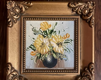 Puede incluir: Una pequeña pintura cuadrada en un marco dorado decorativo con detalles ornamentales en las esquinas. La obra presenta una pintura al óleo de flores amarillas en un jarrón oscuro sobre un fondo claro, con pinceladas visibles y textura de pintura gruesa.