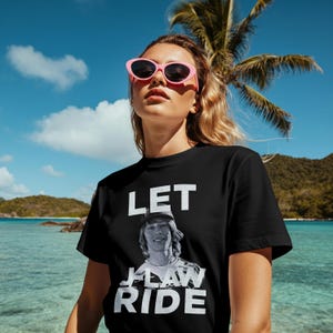 May include: Black t-shirt with the text "LET J-LAW RIDE" in white. The shirt features a black and white image of a person. The background includes a palm tree, blue sky, and ocean.