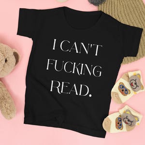 以下が含まれることがあります： 白い文字で「I CAN'T FUCKING READ.」と書かれた黒いTシャツ。シャツは、テディベアのデザインのオフホワイトの靴下と組み合わせています。左側にはぬいぐるみが見えます。