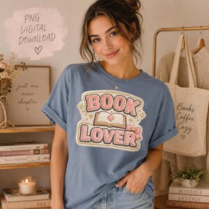 Op de afbeelding: Lichtblauw T-shirt met de tekst "BOOK LOVER" in roze en goud. Het shirt heeft opgerolde mouwen. Een draagtas met de woorden "Books Coffee Cozy" is zichtbaar op de achtergrond. De tekst "PNG DIGITAL DOWNLOAD" is ook aanwezig.