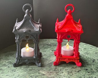 May include: Two decorative lanterns, one black and one red, each with a faux candle inside. The lanterns have a gothic architectural design with a loop at the top for hanging. The lanterns are on a green marble-like surface.